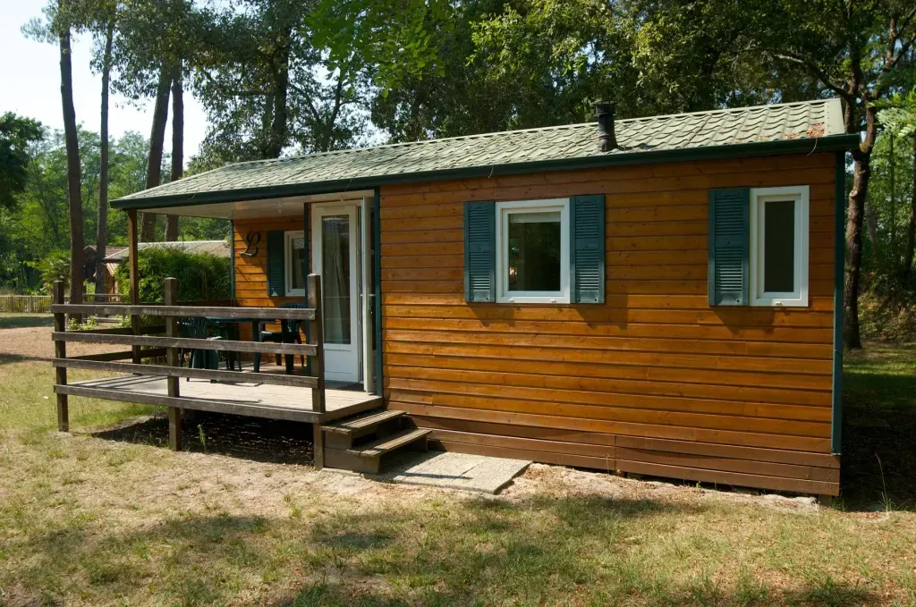 Mobil-home en bois brun avec volets verts et terrasse couverte sur une parcelle arborée