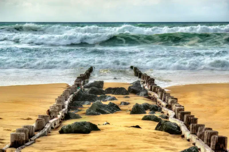 Vagues de l’océan arrivant sur une plage de sable traversée par des pieux en bois et des rochers